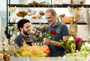 Two florists working together in a flower shop, with an older man holding red roses for a younger colleague surrounded by floral arrangements.
